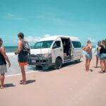 A group of people stand on a sandy beach near a white van that is stuck in the sand, with the ocean and blue sky in the background.