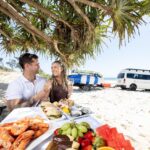 A man and woman sit at a table on a beach, enjoying seafood and fruit, with a van and surfboards parked nearby under a tree.
