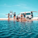 Six people in swimwear pose and jump by a body of water on a sandy beach, with a white van parked in the background under a clear blue sky.