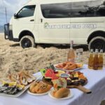 A table with seafood, sandwiches, fruit, drinks, and wine is set up on sand in front of a white "Day Adventure Tours" van parked on a beach under a partly cloudy sky.