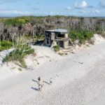 A sandy beach with a few people walking near old concrete bunkers and dense coastal vegetation in the background.