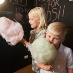 Two young children holding and eating large sticks of cotton candy, standing in front of a chalkboard wall with "fairy floss" written on it.