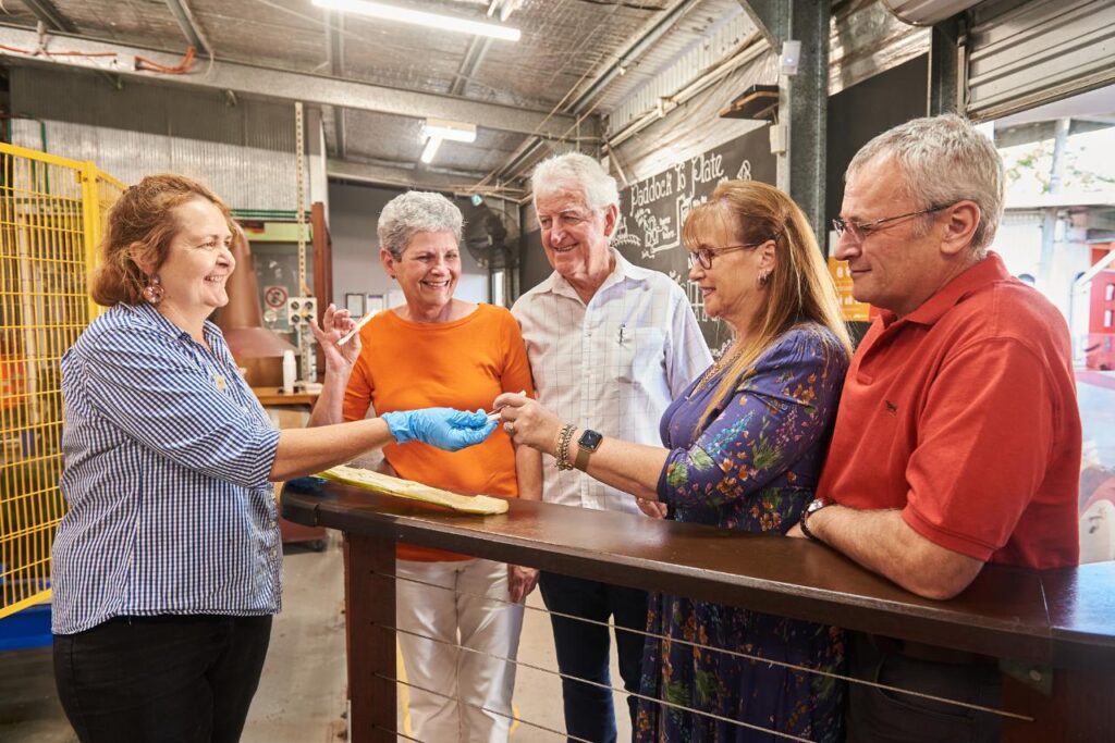 A woman wearing gloves hands an object to a group of four smiling adults standing by a counter in an industrial indoor setting.