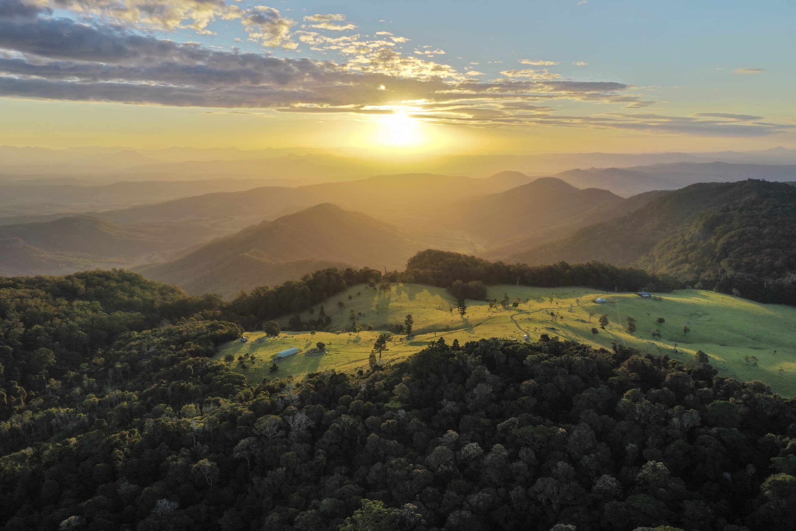 Aerial view of rolling green hills and forested mountains at sunrise, with sunlight casting long shadows across the landscape and a partly cloudy sky above.