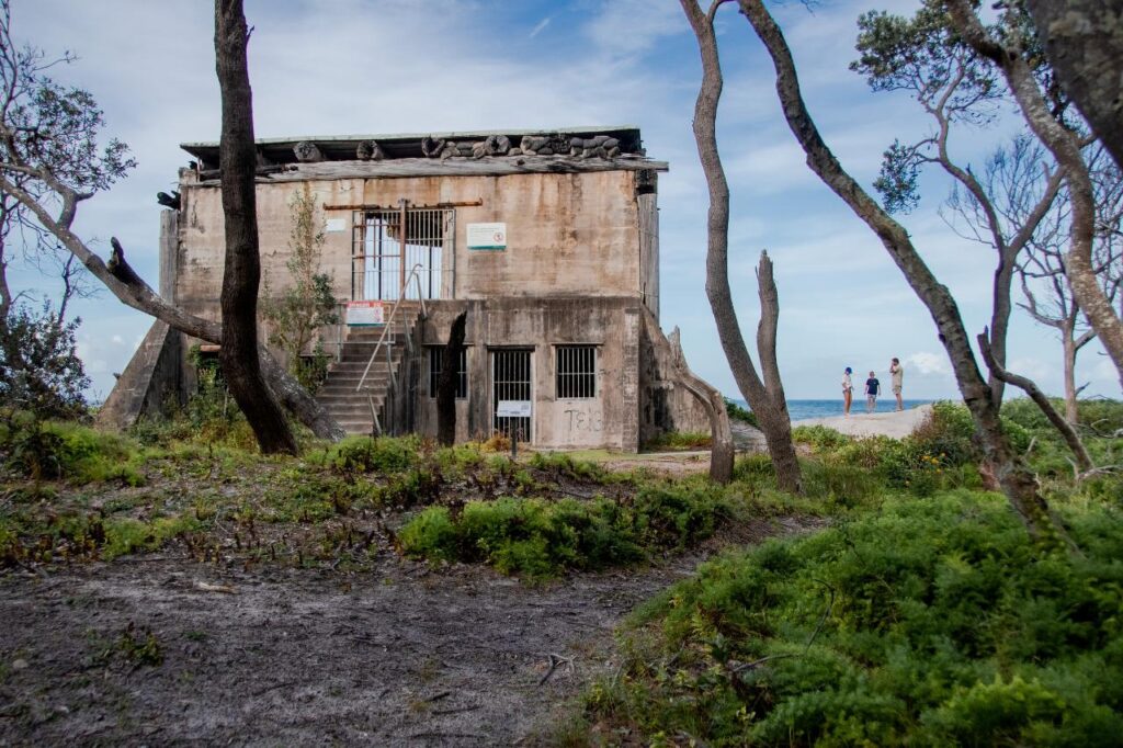 An old concrete building with barred windows and doors stands amid trees and greenery; three people are visible in the distance near the ocean.