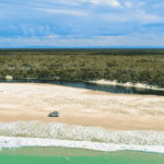 Aerial view of a sandy beach with a parked vehicle near the shoreline, a few people nearby, a narrow river, forest in the background, and waves approaching the shore.