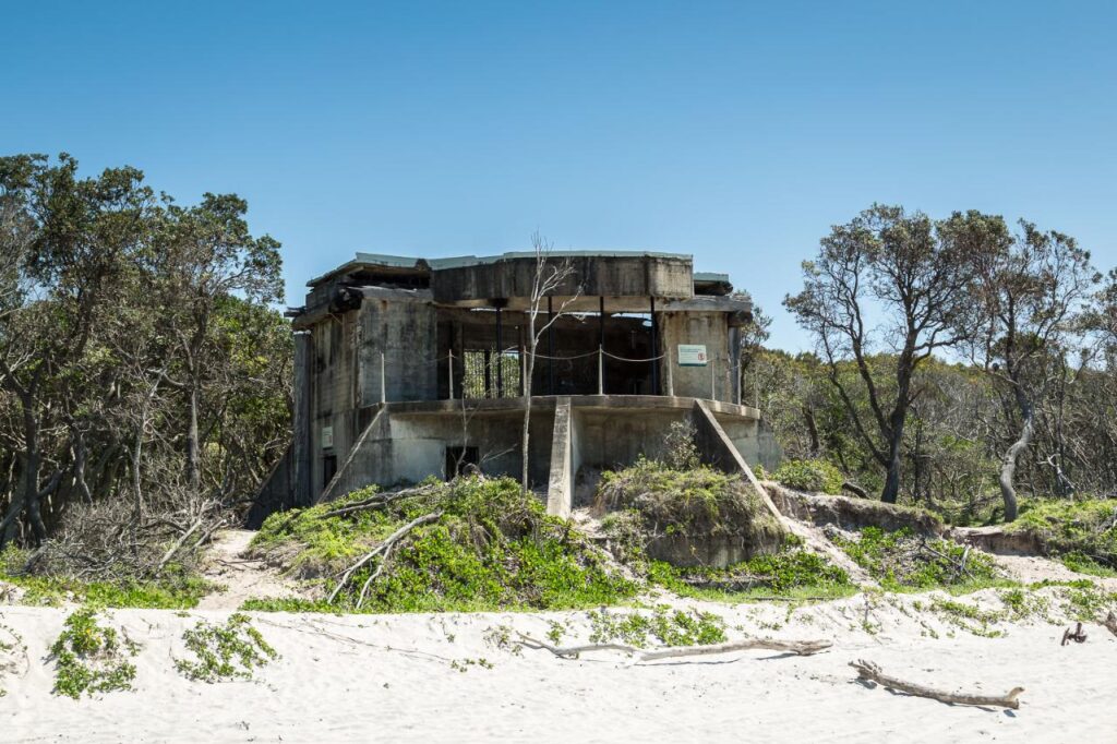 An abandoned concrete bunker stands on a sandy beach, surrounded by sparse vegetation and trees under a clear blue sky.
