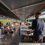 People seated on benches and standing aboard a covered river tour boat, traveling along a scenic river bordered by greenery under a clear sky.