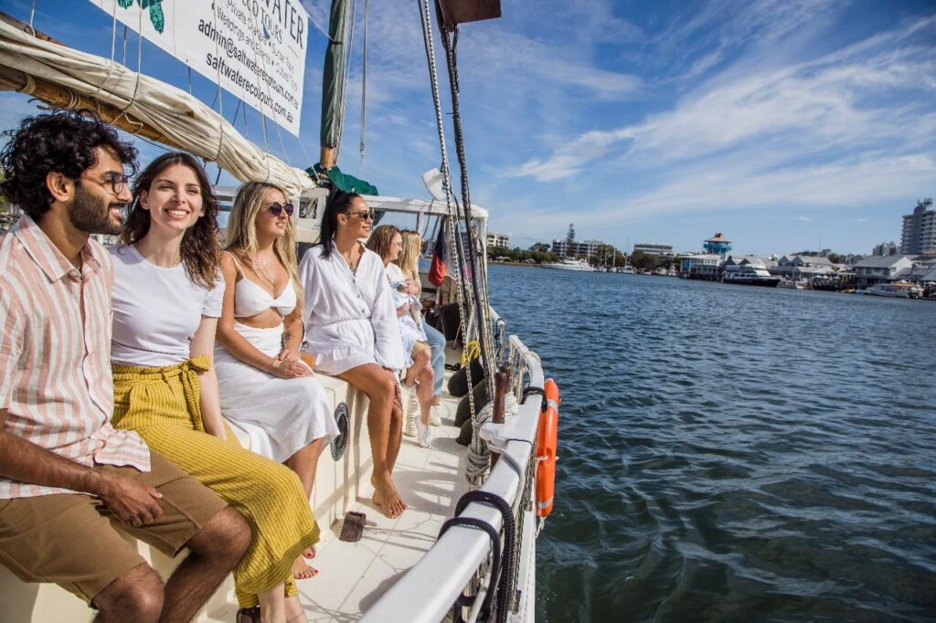 A group of people sits on the edge of a boat, smiling and enjoying a sunny day on the water near a city shoreline.