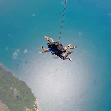 Two people tandem skydiving above a coastline, suspended by a harness, with the ocean and land visible far below.