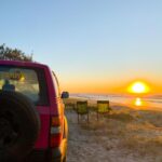 A red SUV parked on sandy ground near two empty camping chairs facing the ocean at sunset, with clear skies and the sun low on the horizon.