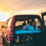 A person lies on a mattress in the back of an SUV parked on a sandy beach at sunset, with the vehicle’s rear and side doors open.