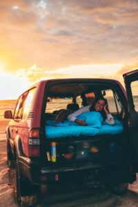 A person lies on a mattress in the back of an SUV parked on a sandy beach at sunset, with the vehicle’s rear and side doors open.