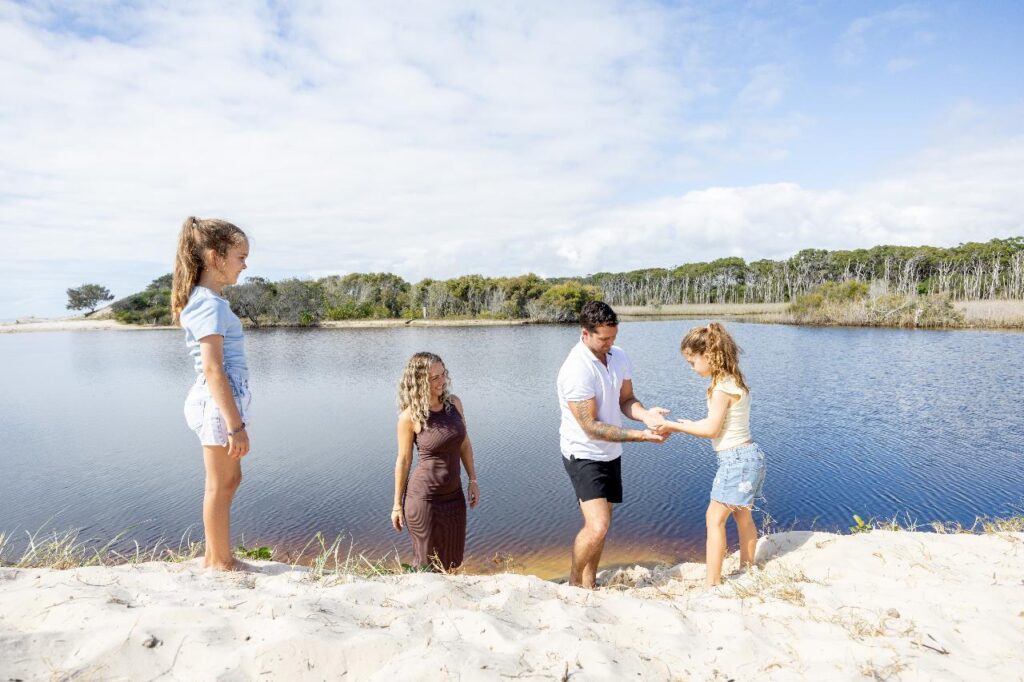 Four people, two adults and two children, stand on sandy ground by a calm lake with trees in the background; one adult helps a child up the slope.