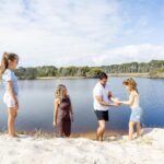 Four people, two adults and two children, stand on sandy ground by a calm lake with trees in the background; one adult helps a child up the slope.