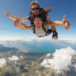 Two people tandem skydiving above clouds and landscape, both smiling and giving thumbs up, with a clear view of land, water, and sky below.
