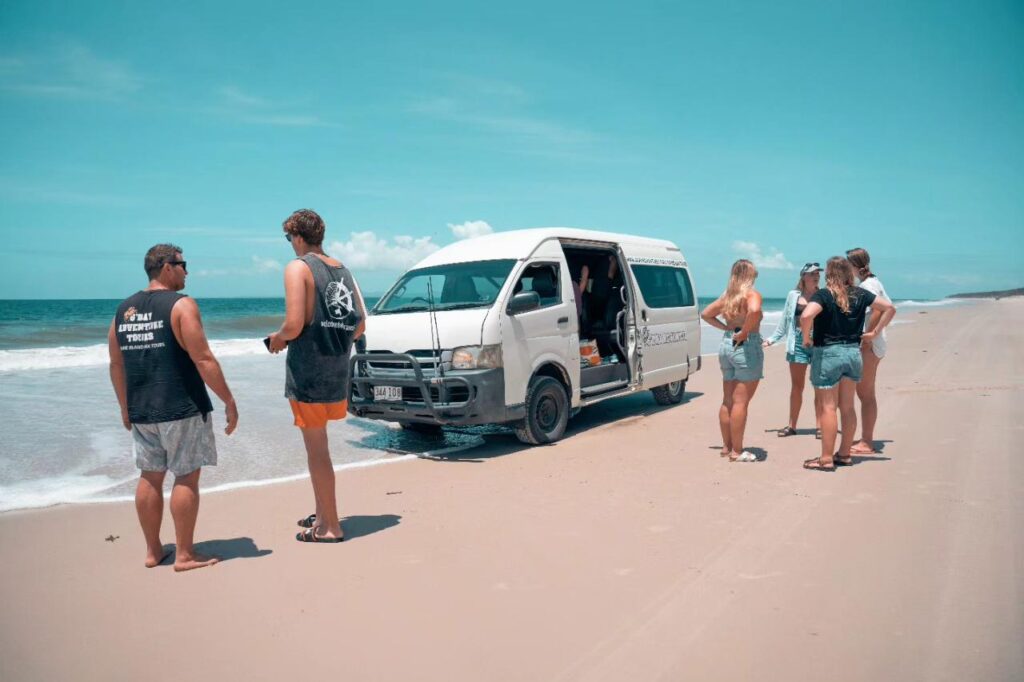 A group of people stand on a sandy beach near a white van with its door open, parked close to the shoreline under a clear blue sky.