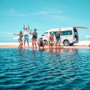 Six people in swimwear pose and jump excitedly by the water on a sandy beach, with a white camper van parked in the background under a clear blue sky.