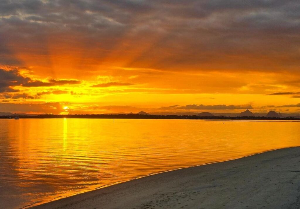 Golden sunset over calm water, with rays of sunlight breaking through clouds and reflecting on the surface near a sandy shoreline.