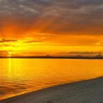Golden sunset over calm water, with rays of sunlight breaking through clouds and reflecting on the surface near a sandy shoreline.