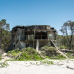 Abandoned concrete bunker with broken windows and overgrown vegetation, situated on a sandy beach with trees in the background under a clear blue sky.