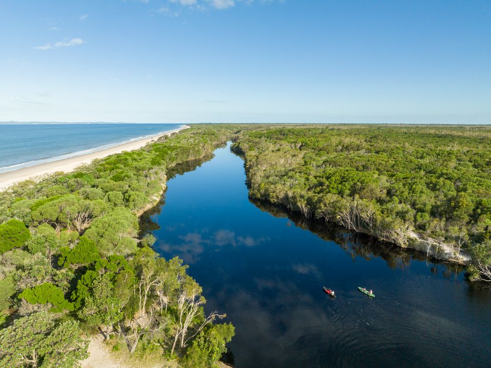Aerial view of a river bordered by dense green forest, with two kayaks on the water and a sandy beach along the coastline under a clear sky.