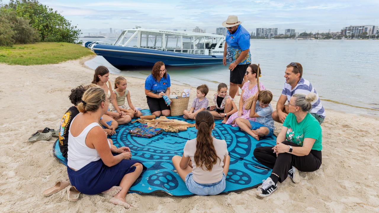 A group of adults and children sit in a circle on a blue patterned blanket on the sand near a body of water, with a boat and city buildings in the background.