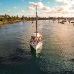 A sailboat with a group of people on board moves through calm water near a marina at sunset, with trees and docks in the background.