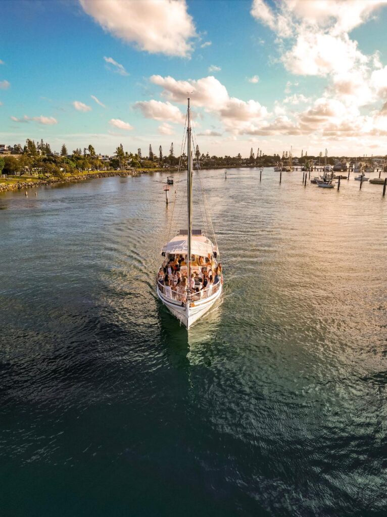 A sailboat with a group of people on board moves through calm water near a marina at sunset, with trees and docks in the background.