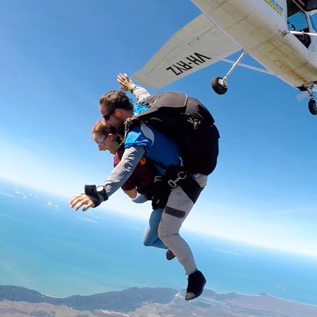 Two people tandem skydiving after jumping from an airplane, with the coastline and ocean visible far below under a clear blue sky.