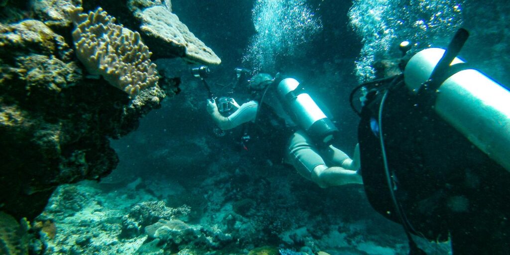 Two scuba divers swim near a coral reef underwater, with diving tanks and gear visible amidst bubbles and marine life.