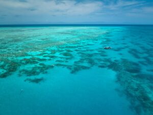 Aerial view of a clear turquoise ocean with patches of coral reefs and a small boat floating on the water under a partly cloudy sky.