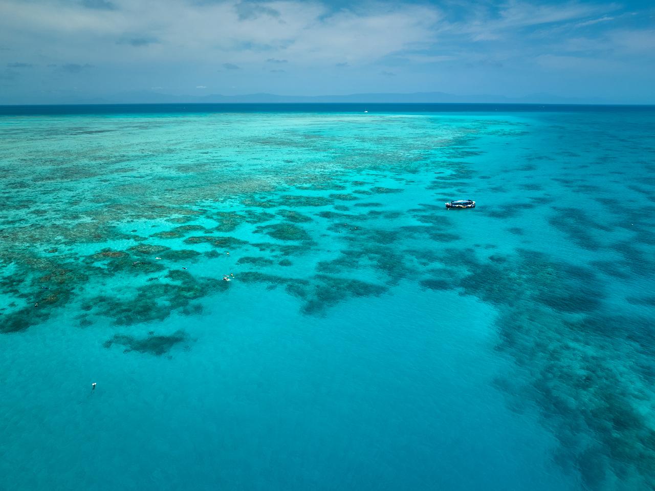 Aerial view of a clear turquoise ocean with patches of coral reefs and a small boat floating on the water under a partly cloudy sky.