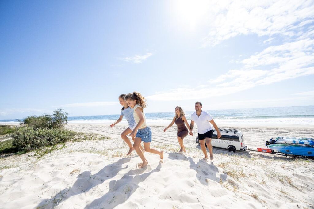 A family of four walks barefoot up a sandy beach dune on a sunny day, with a parked van and surfboards visible in the background near the shoreline.