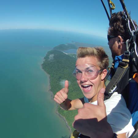 Two people tandem skydiving over a coastline; the person in front is smiling and giving two thumbs up, with land and ocean visible below.