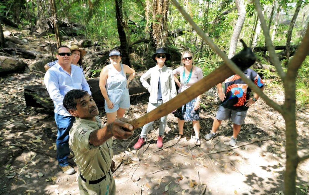 A guide demonstrates how to use a stick on a tree branch while a group of six people watch in a forest setting.