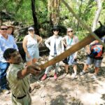 A guide demonstrates how to use a stick on a tree branch while a group of six people watch in a forest setting.