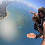 Two people tandem skydiving over a coastal landscape with blue water, shoreline, and clouds visible below them.