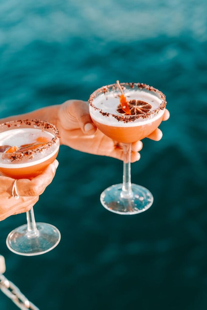 Two hands hold cocktail glasses with frothy drinks, garnished with dried citrus slices and a rim of what appears to be chocolate, against a blue water background.