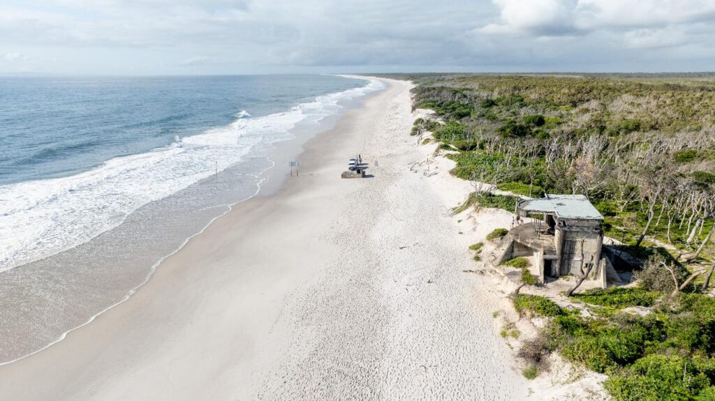 Aerial view of a wide, empty sandy beach bordered by the ocean on one side and green vegetation with a concrete structure on the other.
