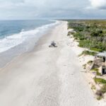 Aerial view of a wide, empty sandy beach bordered by the ocean on one side and green vegetation with a concrete structure on the other.