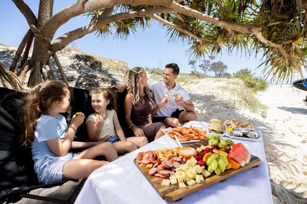 A family of four sits under a tree on a beach, smiling and talking, with a picnic spread of fruit, cheese, and seafood on a table in the foreground.