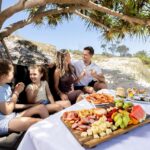 A family of four sits under a tree on a beach, smiling and talking, with a picnic spread of fruit, cheese, and seafood on a table in the foreground.