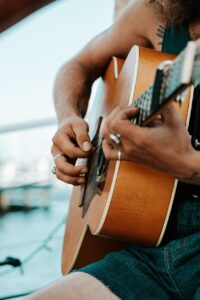 A person wearing rings and sleeveless clothing is playing an acoustic guitar outdoors near the water.