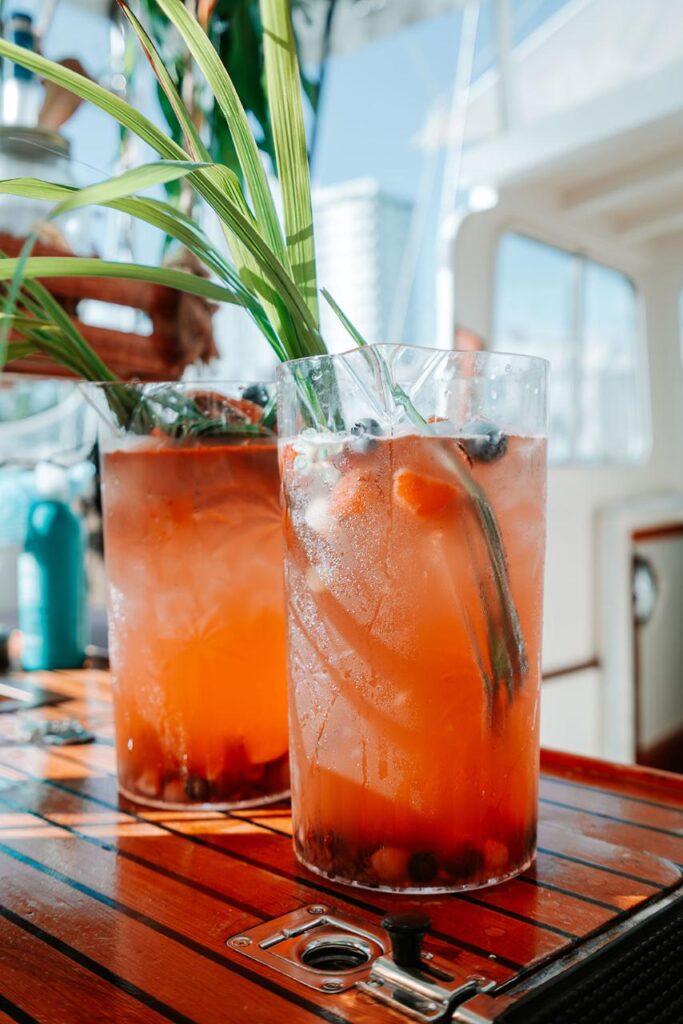 Two tall glasses of iced orange-colored fruit drink with berries and green garnish sit on a wooden table, with a blurred outdoor background.