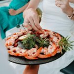 Person in white dress takes a shrimp from a round platter of shrimp garnished with herbs and lemon slices; another person in green dress sits nearby.
