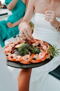 Person in white dress takes a shrimp from a round platter of shrimp garnished with herbs and lemon slices; another person in green dress sits nearby.