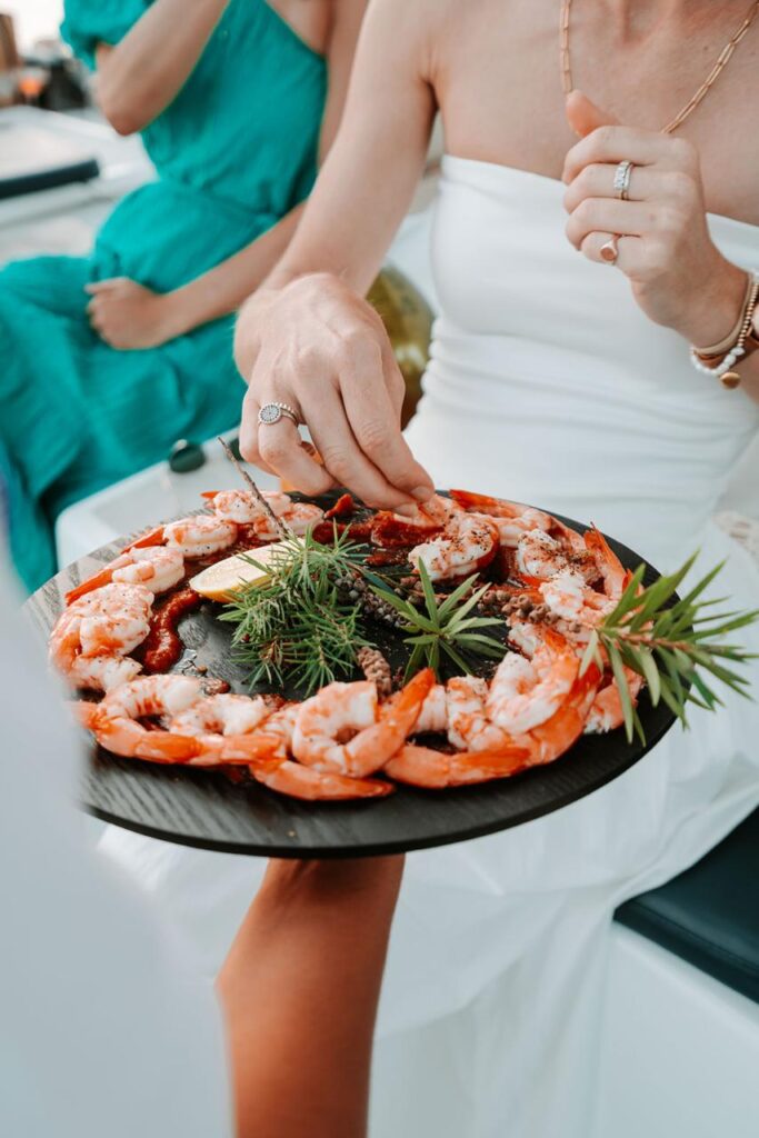 Person in white dress takes a shrimp from a round platter of shrimp garnished with herbs and lemon slices; another person in green dress sits nearby.