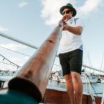 A person standing barefoot on a boat deck plays a didgeridoo, with boats and a marina visible in the background.