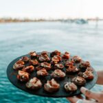 A hand holds a round tray with bite-sized appetizers above blue water, with a blurred shoreline and boats in the background.